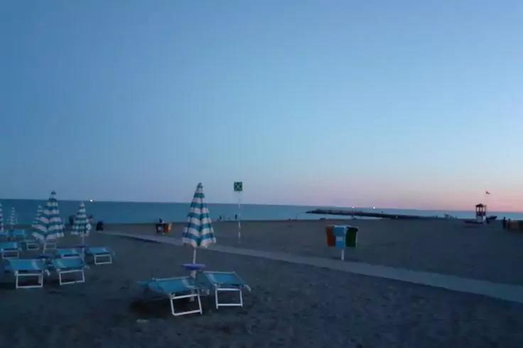 Sandstrand mit leeren Liegest&uuml;hlen und gestreiften Sonnenschirmen, gepflasterter Weg mit M&uuml;llbeh&auml;ltern, ruhiges Meer, Steinmole und kleiner Turm rechts am Horizont, Himmel mit blau-rosa Farbverlauf