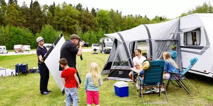 Wohnwagen mit Vorzelt auf einer Wiese, zwei Erwachsene sitzen an einem Campingtisch, ein Erwachsener fotografiert, eine Person h&auml;lt eine Reflektorscheibe, mehrere Kinder stehen auf dem Gras, weitere Wohnwagen und B&auml;ume im Hintergrund