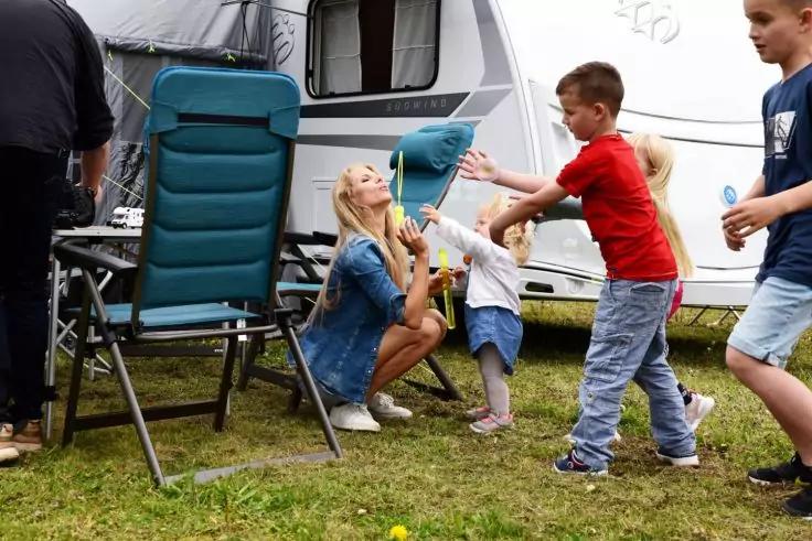 Frau kniet auf Gras vor einem Wohnwagen und h&auml;lt einen Seifenblasenstab; mehrere Kinder strecken die H&auml;nde; Klappst&uuml;hle im Vordergrund
