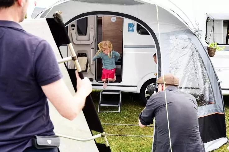 Kind sitzt in ge&ouml;ffneter T&uuml;r eines Wohnwagens; im Vordergrund stellen zwei Personen ein Vorzelt auf; Gras vor dem Wohnwagen