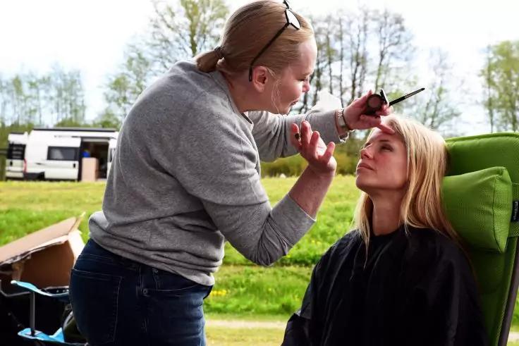 Person tr&auml;gt mit Pinsel Make-up im Gesicht einer sitzenden Frau; Frau mit blonden Haaren auf gr&uuml;nem Stuhl mit schwarzem Umhang; Hintergrund: Wiese und Wohnwagen