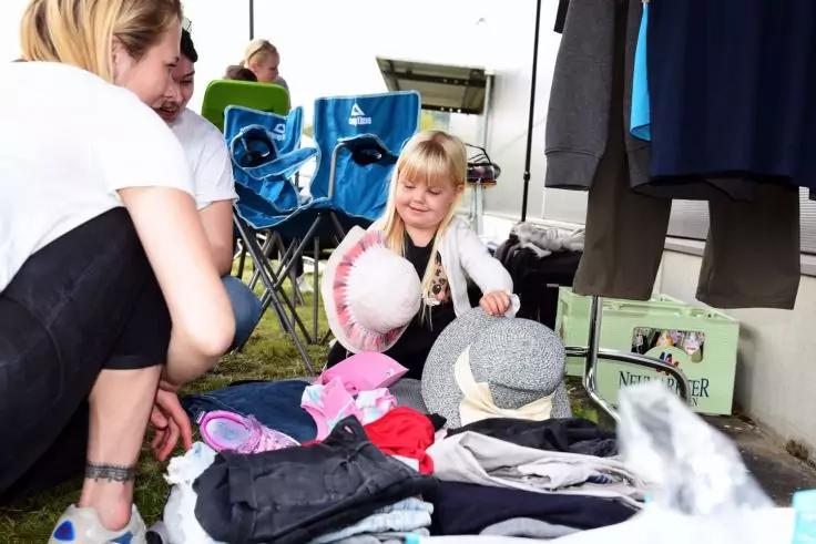 Person mit blonden Haaren sitzt auf dem Boden neben einem Stapel Kleidung und h&auml;lt zwei H&uuml;te; Klappst&uuml;hle und weitere Personen im Hintergrund