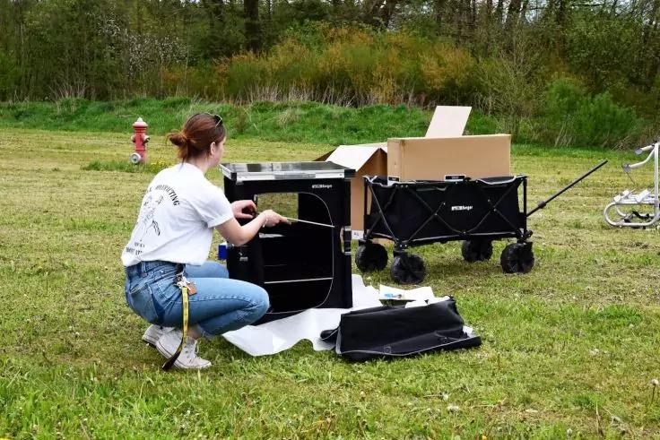 Eine Person kniet auf einer Wiese und greift in eine offene schwarze Transportkiste; auf einer Decke daneben liegen weitere schwarze Kisten und Teile; dahinter ein schwarzer Bollerwagen mit offenem Karton; links ein roter Hydrant; im Hintergrund B&auml;um