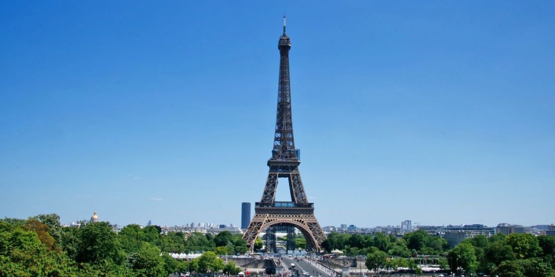 Eiffelturm mittig vor blauem Himmel, im Vordergrund B&auml;ume und Stra&szlig;e