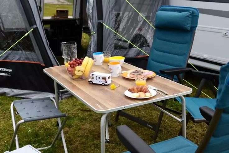 Campingtisch mit Glaskanne, Bechern, Bananen, Weintrauben, Teller mit Croissants, wei&szlig;-schwarzem Gef&auml;&szlig;, blauen Campingst&uuml;hlen, Zelt und Wohnwagen im Hintergrund auf Gras