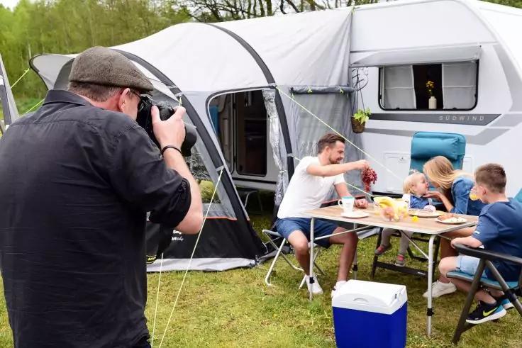Fotograf links mit Kamera. Drei Personen sitzen an einem Campingtisch vor einem Wohnwagen mit angebautem Vorzelt; Teller und Lebensmittel auf dem Tisch, K&uuml;hlbox auf dem Gras