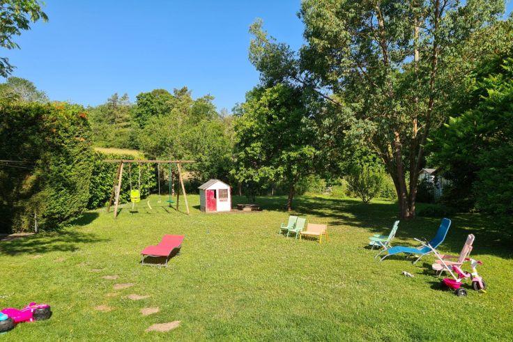 Rasenfl&auml;che mit kleinem wei&szlig;-rosa Spielhaus, Schaukelger&uuml;st mit zwei Schaukeln, mehrere Liegest&uuml;hle und Kinderspielzeug, B&auml;ume und blauer Himmel