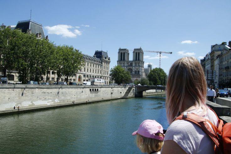 Blick auf einen Fluss mit steinerner Ufermauer und Br&uuml;cke; im Vordergrund die R&uuml;ckenansichten einer Frau und eines Kindes; im Hintergrund eine Kathedrale mit zwei T&uuml;rmen, B&auml;ume, Geb&auml;ude und blauer Himmel