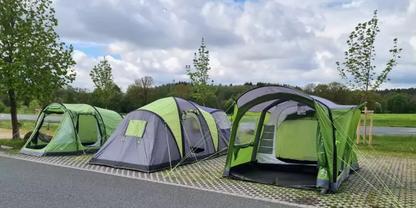 Drei gr&uuml;n-graue Zelte auf gepflasterten Stellpl&auml;tzen neben einer Stra&szlig;e, im Hintergrund B&auml;ume und bew&ouml;lkter Himmel