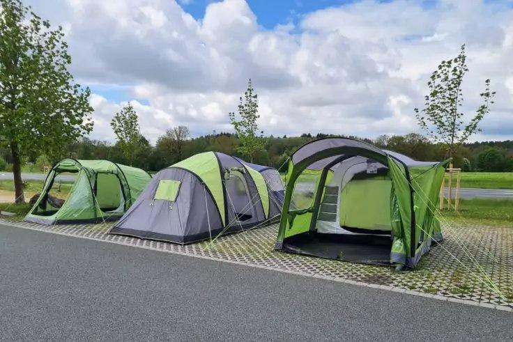Drei gr&uuml;n-graue Zelte auf gepflasterten Stellpl&auml;tzen neben einer Stra&szlig;e, im Hintergrund B&auml;ume und bew&ouml;lkter Himmel