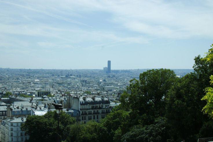 Blick &uuml;ber eine Stadt mit vielen Geb&auml;uden, vorne B&auml;ume, am Horizont ein hoher rechteckiger Turm, blauer Himmel mit Wolken