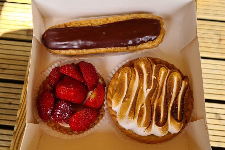 Wei&szlig;e Pappschachtel mit drei Geb&auml;cken: oben ein Eclair mit dunkler Schokoladenglasur, unten links eine Tarte mit halbierten Erdbeeren, unten rechts eine runde Tarte mit goldbraunem Baiser