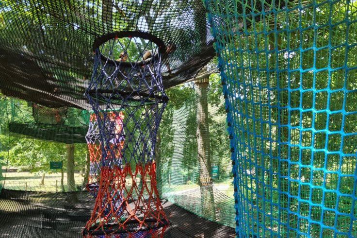 Zylindrisches, h&auml;ngendes Netzgestell in Rot-Orange und Blau, umgeben von blauen und gr&uuml;nen Netzen; B&auml;ume und Holzplattform im Hintergrund