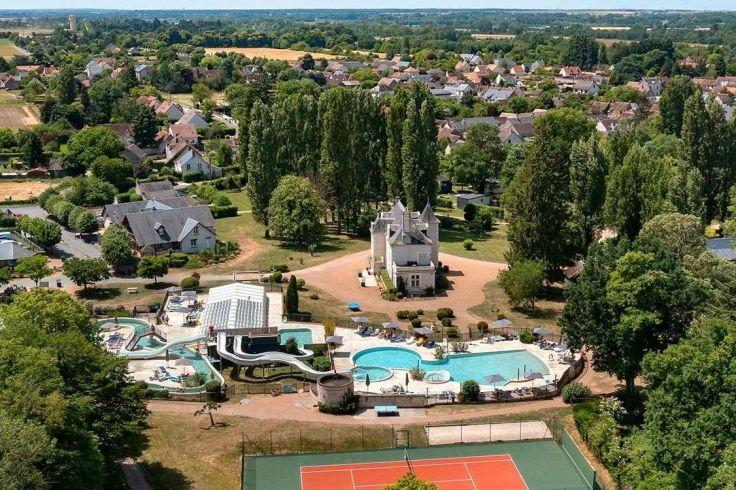 Luftaufnahme von mehreren Au&szlig;enbecken mit Wasserrutsche, einem wei&szlig;en Geb&auml;ude mit Turm, einem Tennisplatz, umgeben von B&auml;umen und Wohnh&auml;usern