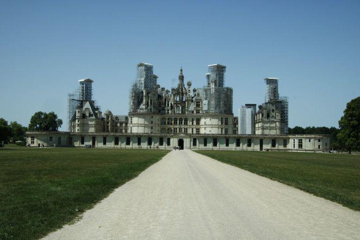 Gerader heller Kiesweg im Vordergrund, f&uuml;hrt zu einem gro&szlig;en symmetrischen Steingeb&auml;ude mit mehreren T&uuml;rmen, Rasenfl&auml;chen links und rechts, klarer blauer Himmel