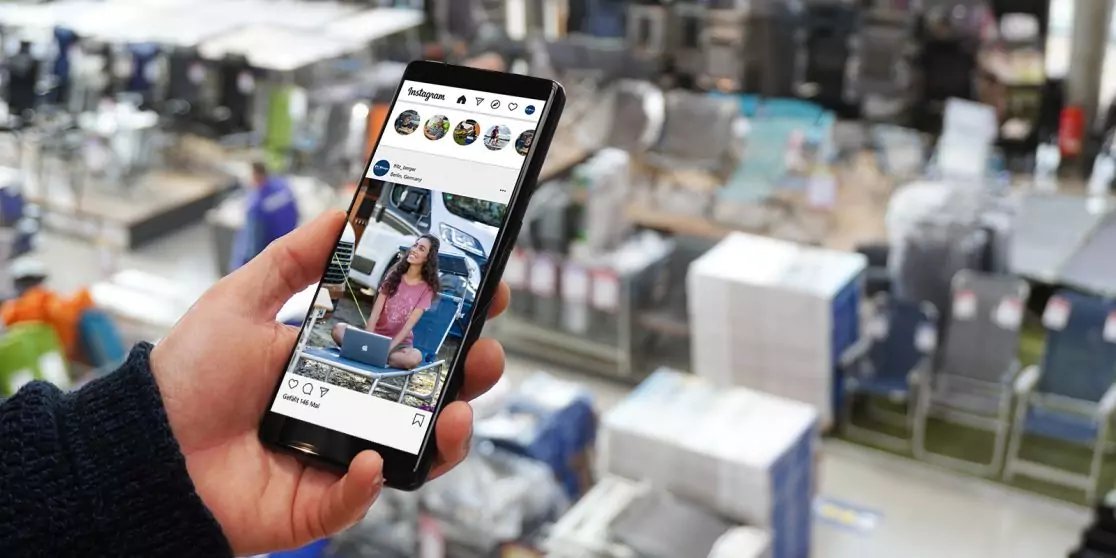 Hand h&auml;lt Smartphone mit offenem Social-Media-Feed; auf dem Bildschirm sind Fotos und Symbole zu sehen; im unscharfen Hintergrund Lagerhalle mit Regalen, Kartons und F&ouml;rderb&auml;ndern