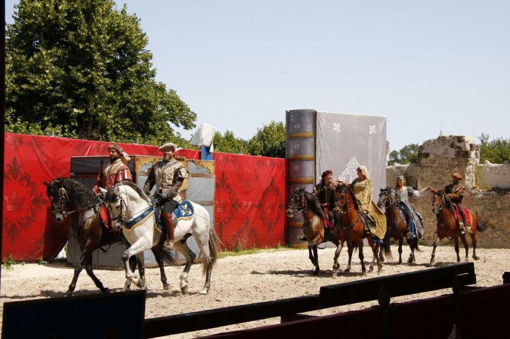 F&uuml;nf Personen in R&uuml;stungen und Kost&uuml;men auf Pferden nebeneinander in einer sandigen Arena; roter dekorativer Vorhang links, B&auml;ume und Ruine im Hintergrund
