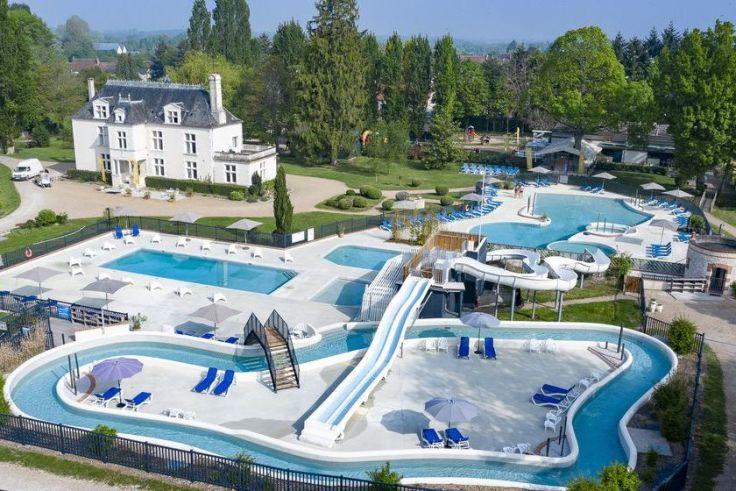 Au&szlig;enbereich mit mehreren verbundenen Schwimmbecken, Wasserrutschen, blauen Liegest&uuml;hlen und einem mehrst&ouml;ckigen wei&szlig;en Geb&auml;ude im Hintergrund, umgeben von B&auml;umen