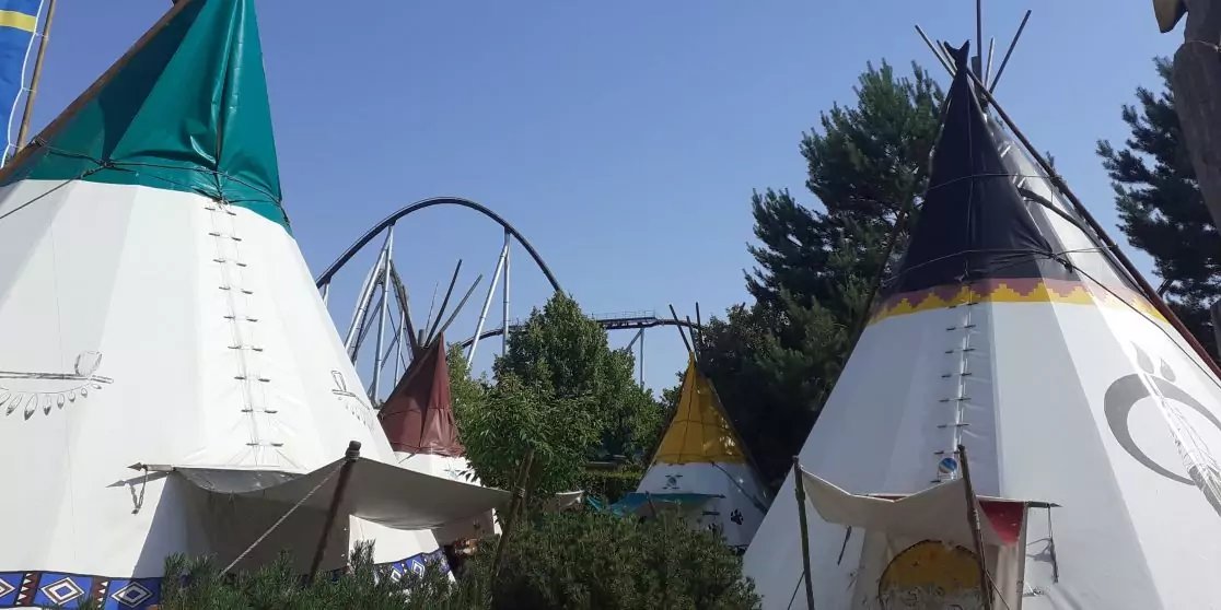 Mehrere Tipis mit Mustern vor B&auml;umen; Achterbahnschienen im Hintergrund; blauer Himmel
