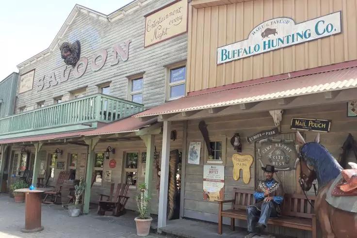 Au&szlig;enansicht einer Western-Geb&auml;udefront mit den Schildern Saloon und Buffalo Hunting Co., Holzveranda, Bank mit sitzender Figur mit Cowboyhut, Pferdefigur rechts