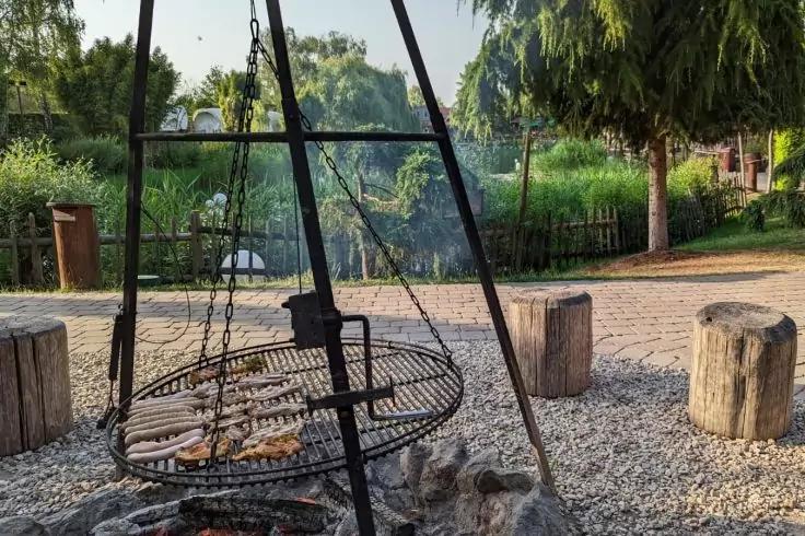 Dreibein-Grill mit rundem, h&auml;ngendem Grillrost &uuml;ber einer Feuerstelle; mehrere Brotscheiben auf dem Rost, aufsteigender Rauch; Kiesboden, Baumstumpfsitzb&auml;nke, B&auml;ume und Holzzaun im Hintergrund