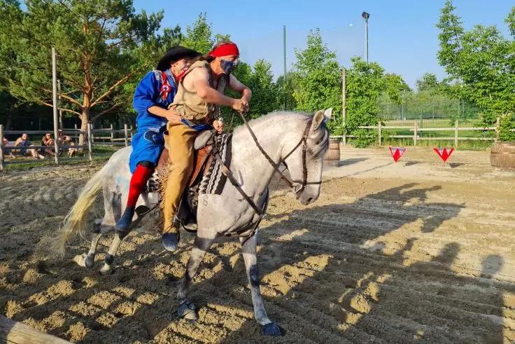 Zwei Personen sitzen auf einem grauen Pferd in einer sandigen Reitbahn; im Hintergrund B&auml;ume, ein Zaun und mehrere Zuschauer