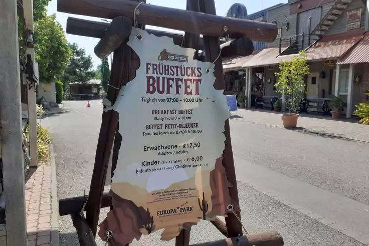 Holz-A‑Schild mit der Aufschrift "FR&Uuml;HST&Uuml;CKS BUFFET" und Preisen auf dem Gehweg; im Hintergrund Stra&szlig;e, H&auml;user und Pflanzk&uuml;bel