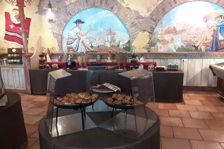 Runde Auslage mit Geb&auml;ck unter transparenten Hauben; im Hintergrund ein Tresen mit Ger&auml;ten und ein gro&szlig;es Wandgem&auml;lde