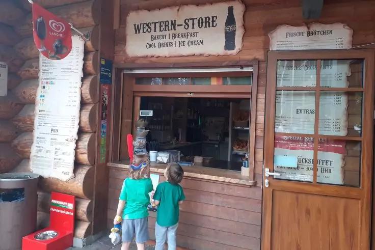 Zwei Personen von hinten in gr&uuml;nen T-Shirts stehen an einem h&ouml;lzernen Kiosk mit ge&ouml;ffnetem Verkaufsfenster; dar&uuml;ber ein Schild &sbquo;Western-Store&lsquo; und links eine Men&uuml;tafel