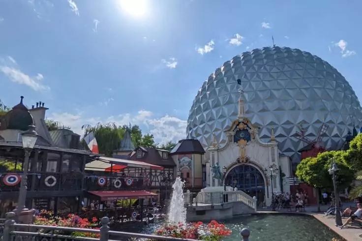 Geod&auml;tische Kuppel rechts, verziertes Bogenportal, Springbrunnen mit Becken und Blumen im Vordergrund, Menschen und traditionelle Geb&auml;ude mit Wimpeln, blauer Himmel mit Sonne und Wolken