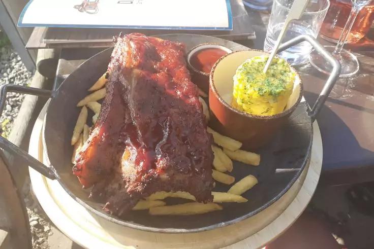 Metallservierplatte mit einem gro&szlig;en gl&auml;nzenden Rippchenst&uuml;ck auf Pommes frites, daneben kleines Sch&auml;lchen mit roter Sauce und kleiner Topf mit gelber Kr&auml;uterbutter; Gl&auml;ser und Speisekarte im Hintergrund