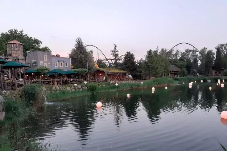 Ufer eines Sees mit kugelf&ouml;rmigen Leuchten auf der Wasseroberfl&auml;che, Geb&auml;uden mit Au&szlig;ensitzbereichen und gr&uuml;nen Sonnenschirmen links, B&auml;umen und zwei geschwungenen Achterbahnschienen im Hintergrund
