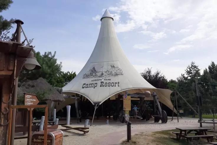 Wei&szlig;es konisches Stoffzelt mit aufgedruckter Illustration und der Schrift Camp Resort, davor Holzsitzb&auml;nke und Picknicktische, Holzwegweiser, Laterne, B&auml;ume und blauer Himmel mit Wolken