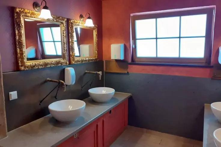 Zwei wei&szlig;e Aufsatzwaschbecken auf grauer Arbeitsplatte, rote Unterschr&auml;nke, zwei goldgerahmte Spiegel mit Lampen, zwei wei&szlig;e Wandspender, Fenster mit unterteilten Scheiben, gefliester Boden