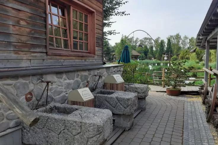 Holzgeb&auml;ude mit Fenster links, gepflasterter Weg, drei grob behauene Steinb&auml;nke, Pflanzk&uuml;bel, geschlossener gr&uuml;ner Sonnenschirm, Holzzaun, Wasser mit wei&szlig;en Bojen und kleine Bogenbr&uuml;cke im Hintergrund, B&auml;ume und Str&auml;ucher