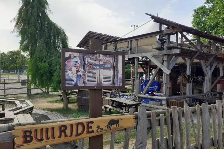 Holzgel&auml;nder und Schild Bullride mit Stiersilhouette vor einer &uuml;berdachten Holzstruktur, einer Plakattafel, B&auml;nken und B&auml;umen