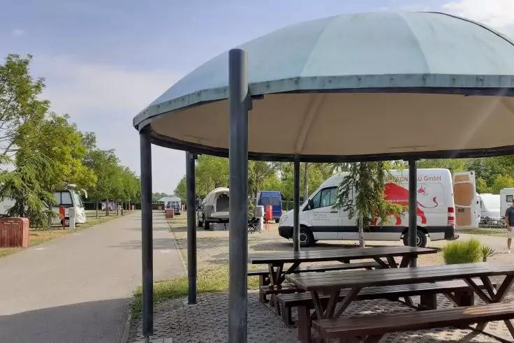 Runder Pavillon mit B&auml;nken und Picknicktischen; im Hintergrund mehrere Wohnmobile entlang einer gepflasterten Stra&szlig;e, B&auml;ume und teilweise bew&ouml;lkter Himmel
