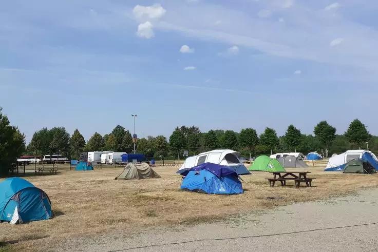 Mehrere kleine Zelte verschiedener Farben auf trockenem, braunem Gras, Picknicktische, B&auml;ume und Wohnwagen im Hintergrund, blauer Himmel mit einzelnen Wolken, gepflasterter Weg im Vordergrund