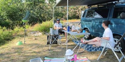 Zwei Personen sitzen auf Campingst&uuml;hlen unter einem Sonnendach neben einem Campingbus; Klapptisch, Decke, Sonnenhut und B&auml;ume im Hintergrund