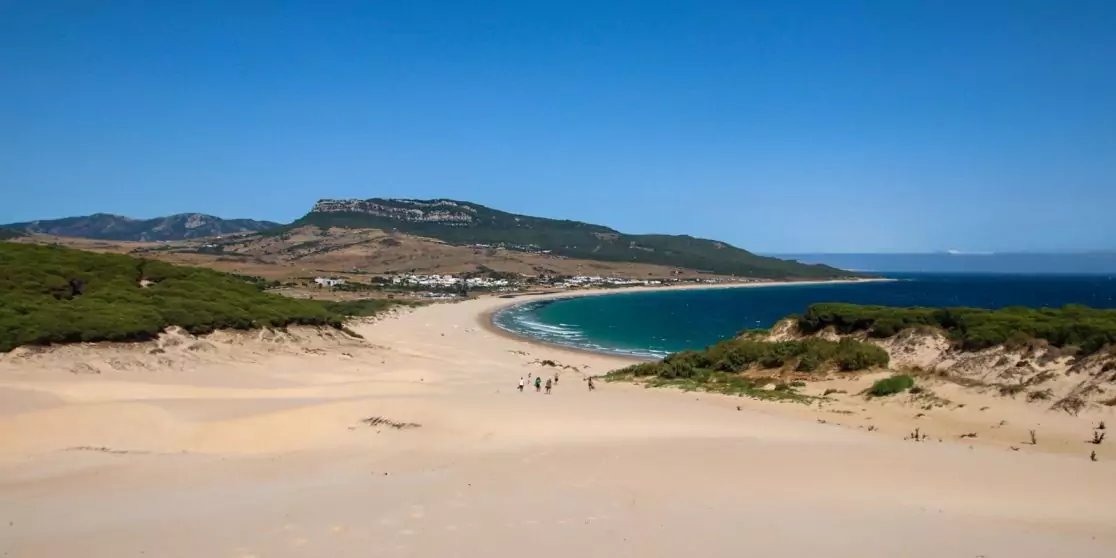 Weiter Sandstrand mit D&uuml;nen im Vordergrund, wenige Personen am Wasser, geschwungene Bucht mit t&uuml;rkisblauem Meer, H&auml;user am Fu&szlig; gr&uuml;ner H&uuml;gel und klarer blauer Himmel