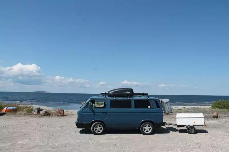 Blauer Transporter mit Dachbox und kleinem wei&szlig;en Anh&auml;nger auf kiesigem Strand; Meer, Horizont und blauer Himmel mit vereinzelten Wolken im Hintergrund; zwei bunte Kajaks links