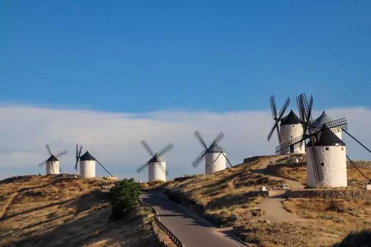 Mehrere wei&szlig;e Windm&uuml;hlen auf einem trockenen H&uuml;gel, ein Weg f&uuml;hrt den Hang hinauf, blauer Himmel mit vereinzelten Wolken