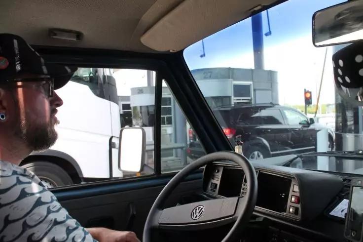 Innenansicht eines Volkswagen: Lenkrad mit VW-Logo, Innenspiegel, Fahrer mit Kappe und Bart; drau&szlig;en Zapfs&auml;ulen, wei&szlig;er Bus links und schwarzer SUV rechts