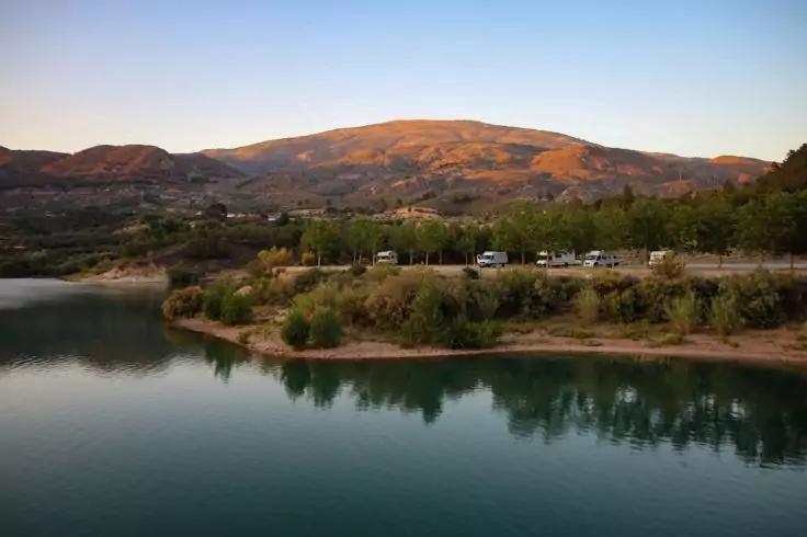 Ufer eines ruhigen Sees mit Spiegelung, B&uuml;schen und B&auml;umen, geparkte Wohnmobile am Ufer, H&uuml;gel im Hintergrund unter klarem Himmel