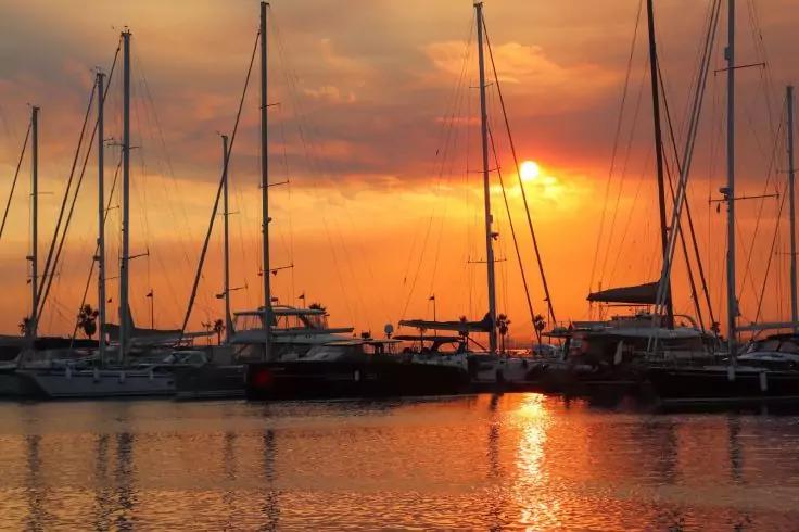 Mehrere Segelboote und Yachten in einem Hafen, orangefarbener Himmel, Sonne teilweise von Wolken verdeckt, Spiegelung im Wasser