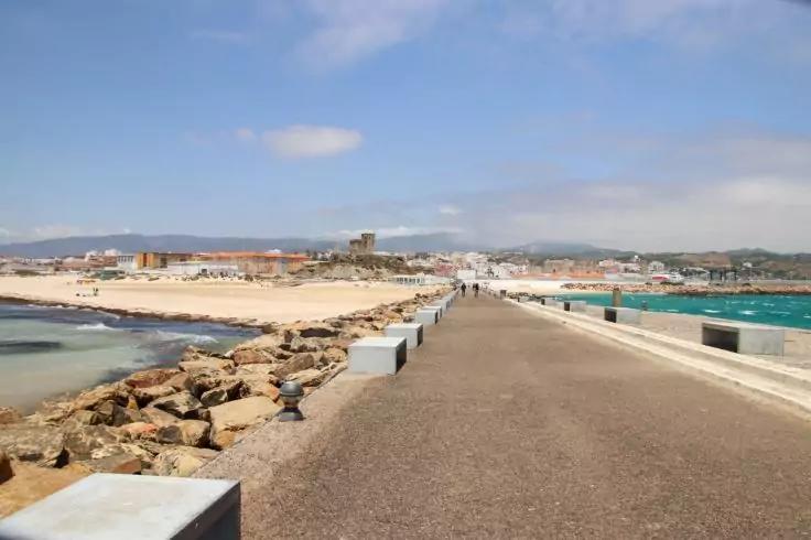 Breite Betonmole mit niedrigen Blockw&auml;nden, rechts t&uuml;rkisfarbenes Meer, links Felsen und Sandstrand, im Hintergrund Geb&auml;ude und blauer Himmel mit Wolken
