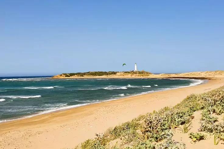 Sandstrand mit D&uuml;nen und Strandvegetation, Wellen im Meer und ein wei&szlig;er Leuchtturm auf einer Landzunge unter blauem Himmel