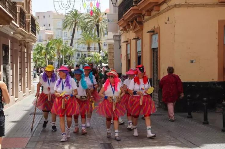 Gruppe von Personen in bunten Kost&uuml;men mit roten R&ouml;cken, wei&szlig;en Socken und Sportschuhen, einige mit H&uuml;ten, gehend auf einer gepflasterten Fu&szlig;g&auml;ngerzone zwischen Geb&auml;uden; Palmen im Hintergrund