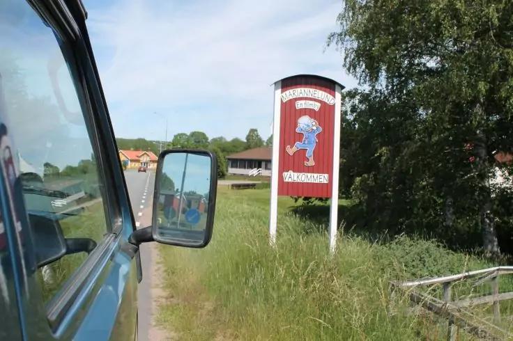 Blick aus blauem Fahrzeug: linker Au&szlig;enspiegel, Stra&szlig;e, grasbewachsene B&ouml;schung, rotes Schild auf zwei wei&szlig;en Pfosten mit blauem Figurenmotiv und Text, H&auml;user und B&auml;ume im Hintergrund, bew&ouml;lkter Himmel