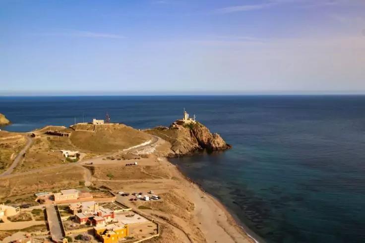 K&uuml;stenlandschaft mit Sandstrand, Felsklippe mit Leuchtturm, mehrere niedrige Geb&auml;ude, ruhiges Meer und blauer Himmel